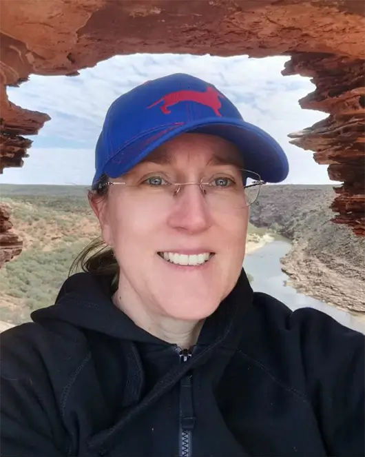 Vicki McMurray wearing glasses and a blue cap smiles for a selfie near a natural rock formation with a river and landscape visible in the background